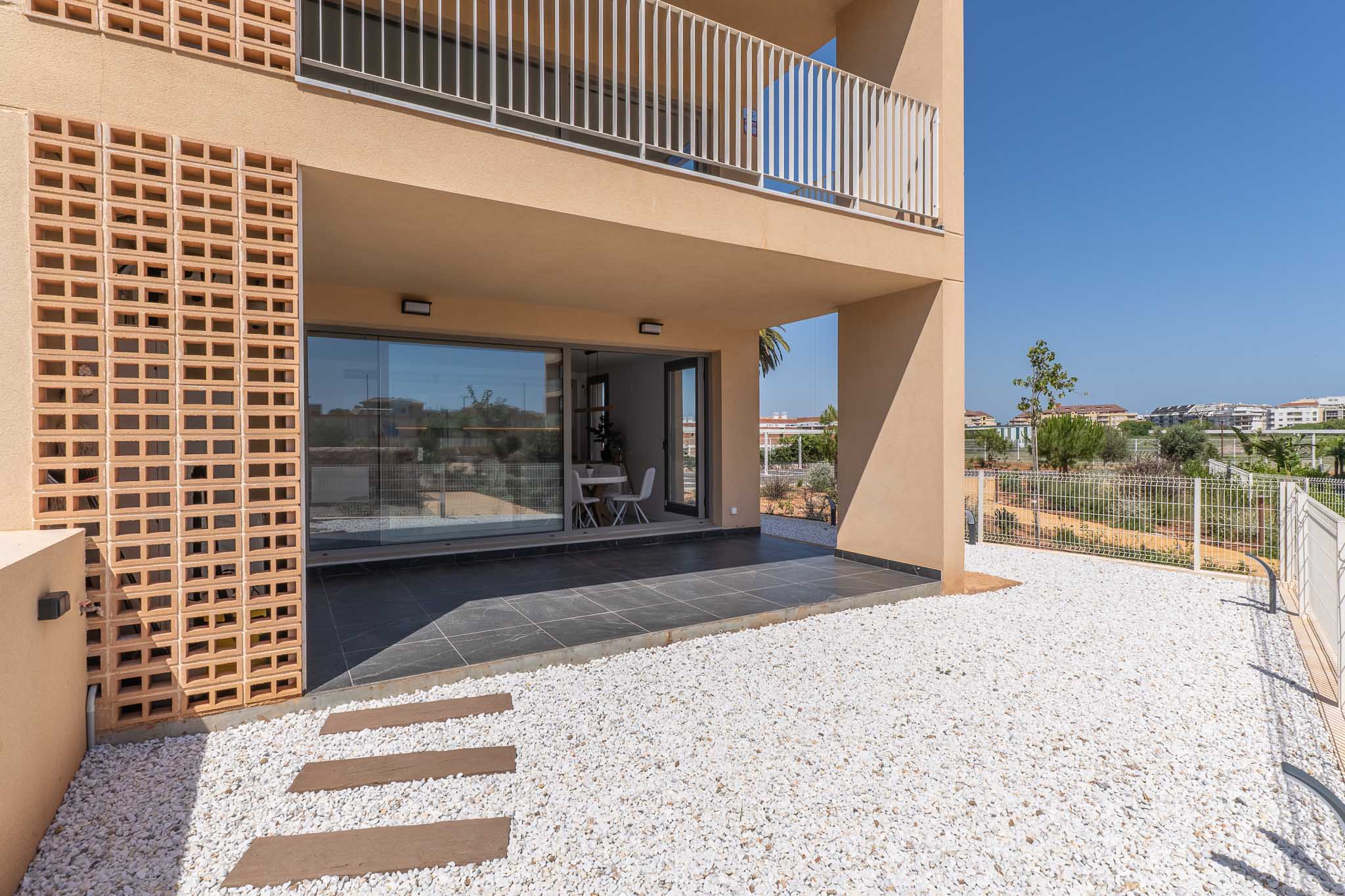 Terraza cubierta de planta baja con baldosas oscuras, conectada a un jardín privado de grava blanca. Grandes puertas de cristal revelan un luminoso comedor interior, junto a una moderna fachada con celosía de ladrillo.