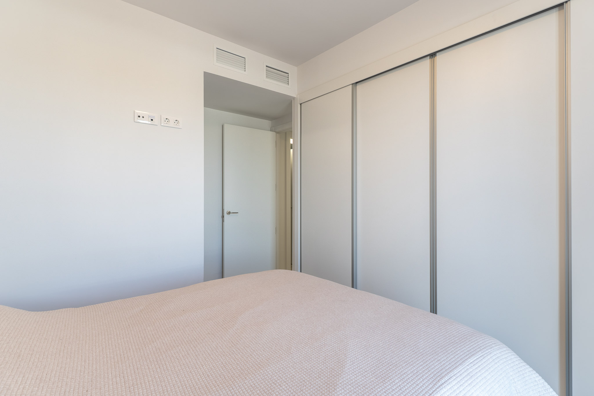 Vista de dormitorio con un amplio armario empotrado de puertas correderas blancas. La pared presenta tomas eléctricas modernas y rejillas de ventilación integradas sobre la puerta adyacente.