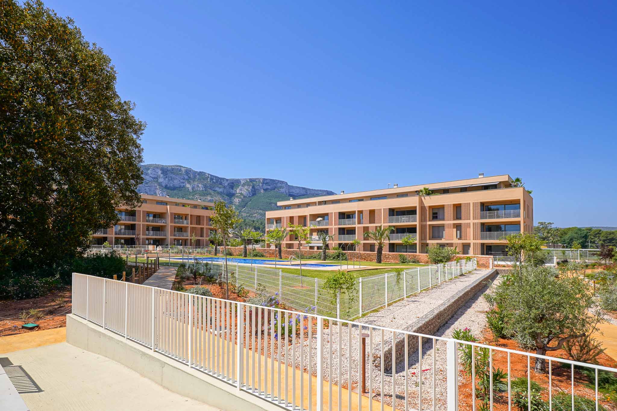 Vista exterior del residencial moderno con dos edificios de fachadas claras, piscina comunitaria y jardines. Al fondo, se aprecian las montañas bajo un cielo despejado.