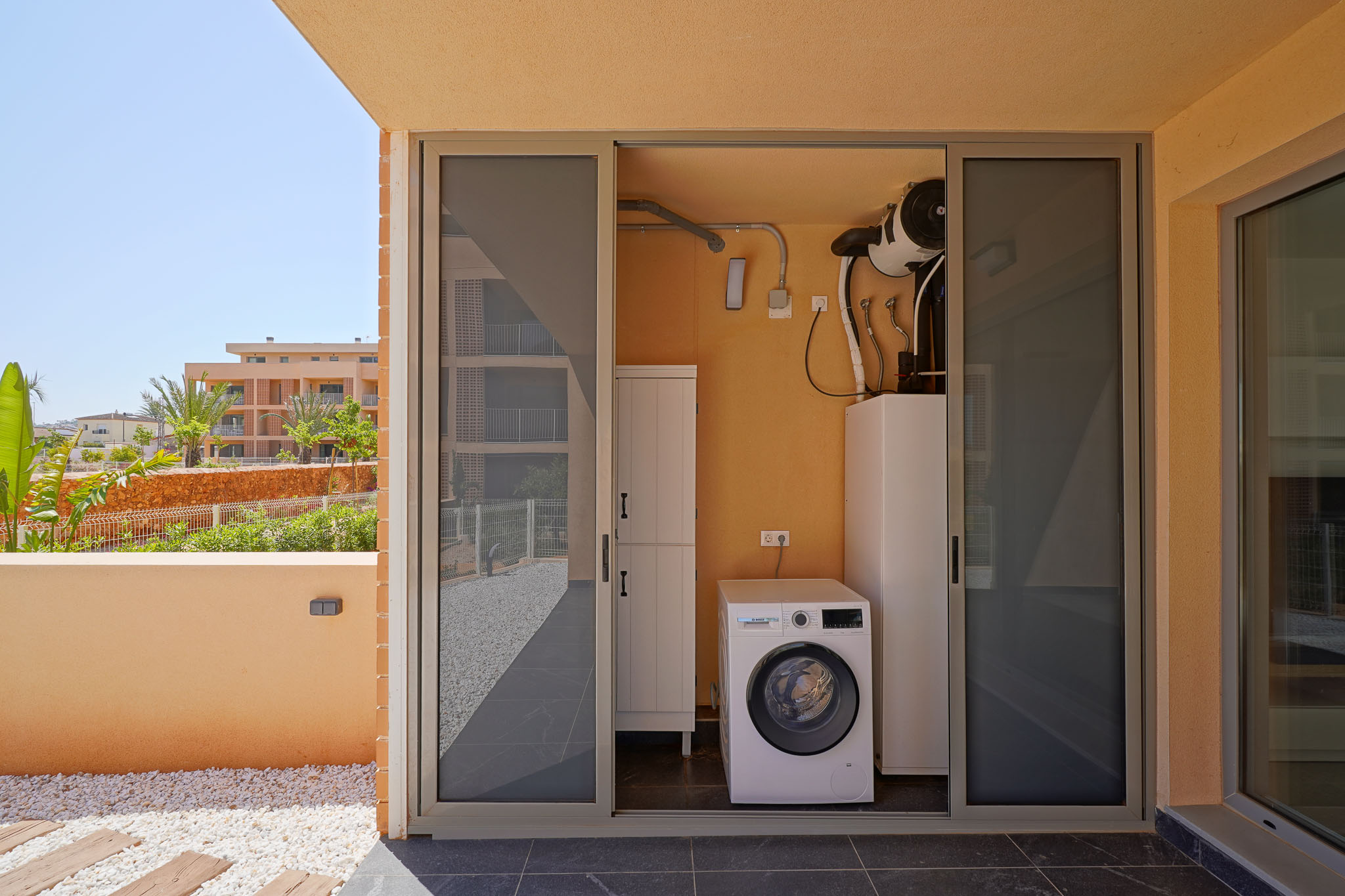 Zona de lavandería exterior con lavadora Bosch y sistema de aerotermia, accesible desde una terraza con suelo de gres, guijarros y vistas al jardín.