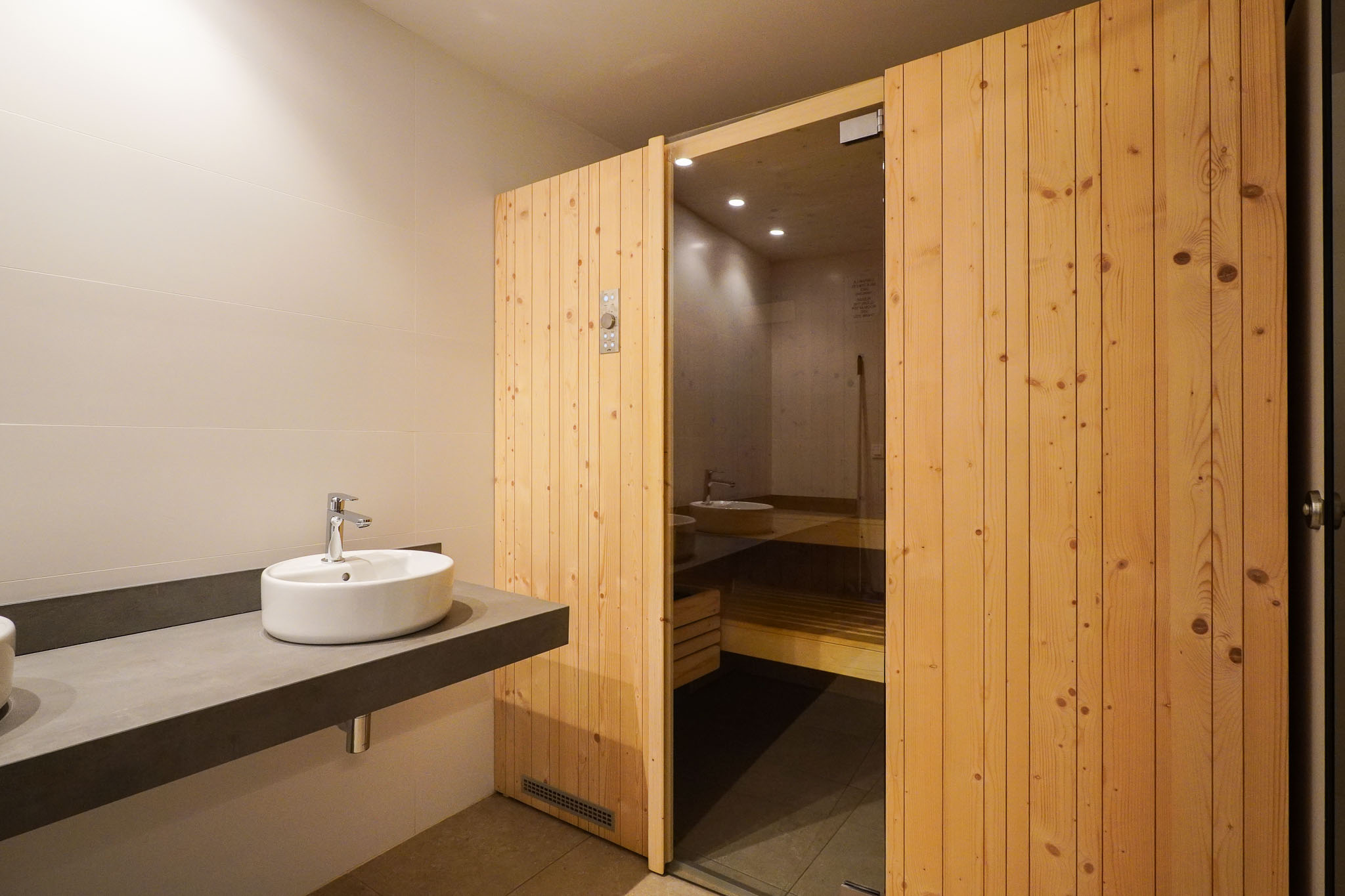 Zona de vestuario con un elegante lavabo sobre encimera flotante de diseño y la entrada a la sauna, con puerta de madera y cristal, parte del centro social.