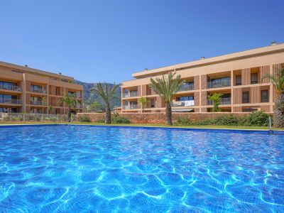 Amplia piscina comunitaria con mosaicos azules brillantes, enmarcada por edificios residenciales modernos con balcones y un telón de fondo montañoso bajo un cielo despejado.