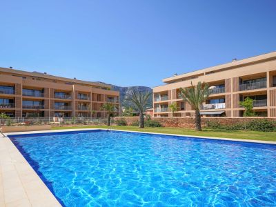 Amplia piscina comunitaria con mosaicos azules, rodeada de césped y edificios residenciales modernos con balcones. Al fondo, se aprecian palmeras y montañas.