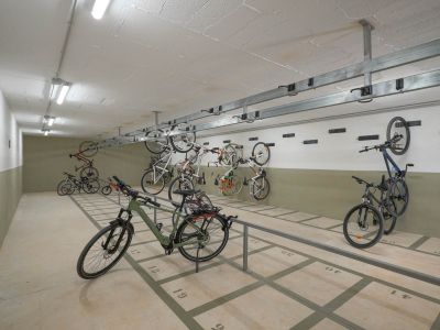 Amplia zona de aparcamiento comunitario para bicicletas con plazas numeradas en el suelo y soportes de pared. Iluminado con luces fluorescentes, ofrece almacenamiento seguro en el complejo residencial.