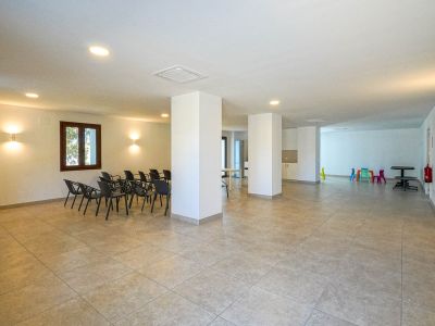 Amplio centro social con suelo cerámico, pilares estructurales y luminarias empotradas. Incluye zona de sillas para reuniones y un rincón infantil con mobiliario colorido.