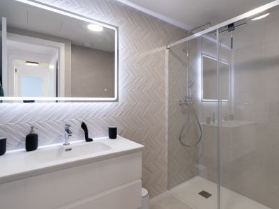 Baño moderno con lavabo blanco, espejo retroiluminado y azulejos en patrón espiga. La ducha con mampara de cristal y grifería cromada complementa el diseño elegante.