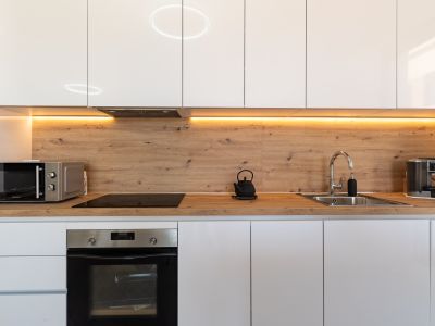 Cocina moderna con armarios blancos brillantes, encimera y salpicadero de efecto madera, e iluminación LED bajo los gabinetes. Equipada con horno, microondas y placa de inducción.