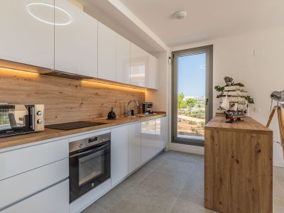 Cocina moderna con armarios blancos brillantes, encimera y salpicadero efecto madera. Equipada con electrodomésticos integrados, iluminación LED y ventana con vistas al exterior.