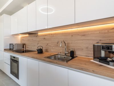 Cocina moderna con mobiliario blanco brillante, encimera y salpicadero de madera clara. Equipada con horno integrado, placa de inducción y fregadero de acero, realzada por iluminación LED bajo los armarios.