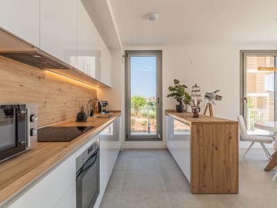 Cocina moderna de diseño abierto con muebles blancos brillantes, encimeras y salpicadero de efecto madera, e iluminación LED bajo los armarios. Incluye electrodomésticos integrados y acceso a la terraza.