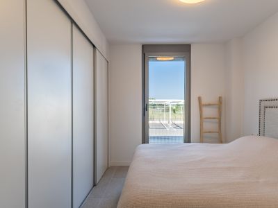 Dormitorio luminoso con armario empotrado de puertas correderas blancas, suelo porcelánico y acceso directo al exterior a través de una puerta acristalada. Cama con cabecero tapizado.