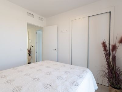 Dormitorio luminoso con cama de edredón blanco y detalles bordados. Incluye armario empotrado con puertas correderas blancas y climatización por conductos. Un toque decorativo con planta ornamental.