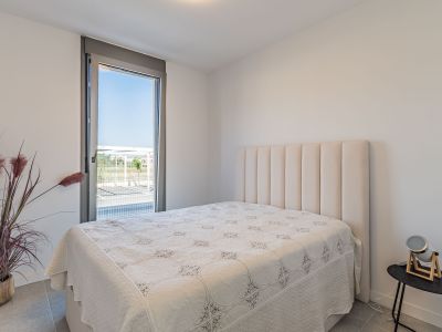 Dormitorio luminoso con cama y cabecero acolchado beige. Gran ventanal de suelo a techo con vistas exteriores y suelo de gres gris. Planta decorativa y mesita auxiliar moderna.