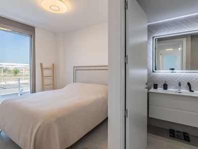 Dormitorio principal con cama de cabecero tapizado y salida directa a una terraza exterior. Se aprecia un baño en suite con lavabo flotante y espejo retroiluminado.