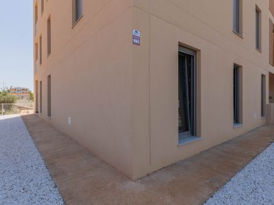 Exterior de edificio moderno con fachada de estuco beige claro y ventanas de marcos oscuros. El suelo presenta un camino de hormigón texturizado junto a un área de gravilla blanca, con alarma visible.