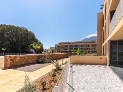 Jardín privado en planta baja con grava blanca, delimitado por una valla baja y una pared de piedra. Se aprecian senderos y vegetación joven, con vistas a edificios modernos y montañas.