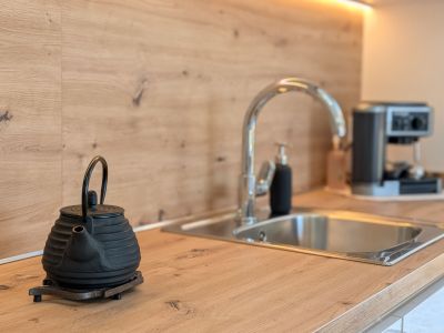 Moderna encimera de cocina con acabado de madera clara y salpicadero a juego. Fregadero de acero inoxidable, grifo cromado y luz LED bajo mueble. Tetera de hierro fundido en primer plano.