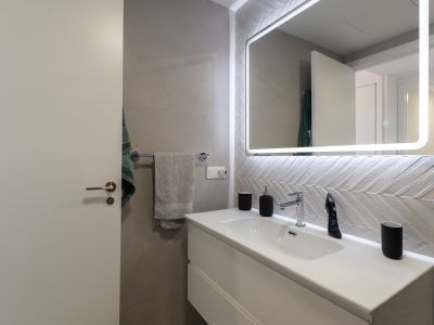 Moderno baño con lavabo flotante blanco, grifería cromada y espejo retroiluminado. Destacan los azulejos texturizados con patrón de espiga en la pared.