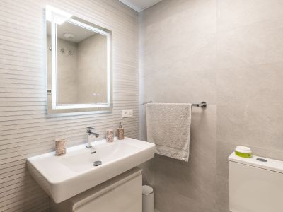 Moderno tocador de baño con lavabo rectangular blanco, grifo mural, espejo iluminado y azulejos de pared grises texturizados.
