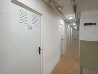 Pasillo de trasteros con puertas blancas numeradas y rejillas de ventilación. El techo muestra conductos de ventilación expuestos e iluminación empotrada, sobre un suelo de hormigón.