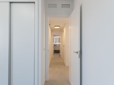 Pasillo luminoso con armario empotrado blanco y puerta abierta. El suelo de baldosas claras conduce a un baño moderno con espejo retroiluminado y mueble de diseño. Rejillas de ventilación en el techo.