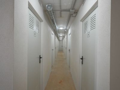 Pasillo utilitario a trasteros individuales, con puertas blancas numeradas y rejillas de ventilación. Techo de hormigón visto con conductos industriales e iluminación empotrada, junto a paredes texturizadas.