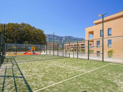 Pista de pádel de césped artificial verde, con vallas y postes de iluminación, junto a un parque infantil y edificios residenciales modernos con vistas a la montaña.