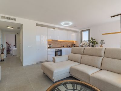 Salón-cocina diáfano con sofá seccional beige, cocina moderna de armarios blancos y encimera de madera clara. Suelo de gres y acceso a exterior.