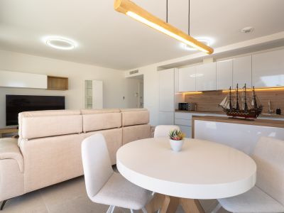 Salón-comedor de planta abierta con cocina integrada. Destaca el sofá beige, mesa ovalada blanca y cocina moderna con muebles lacados blancos, salpicadero de madera y luz ambiental.