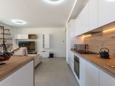 Salón-comedor diáfano con cocina abierta de diseño moderno. Destacan los muebles blancos lacados, encimeras de efecto madera clara y electrodomésticos integrados, complementados con iluminación LED y suelo cerámico gris.