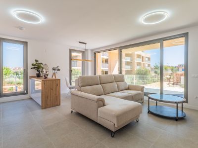 Salón-comedor diáfano con sofá seccional beige y mesa de centro ovalada. Grandes ventanales conectan con el exterior, ofreciendo luz natural y vistas residenciales. Cocina abierta con barra de madera.