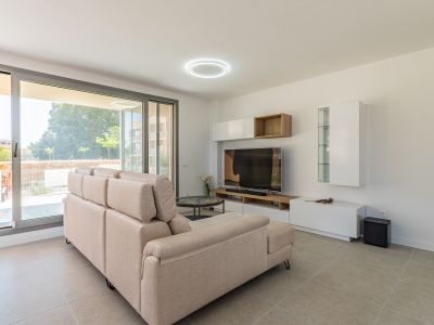 Salón luminoso con sofá seccional beige, suelo de gres claro y amplios ventanales correderos que dan al exterior. Mueble de TV moderno con estantes blancos y de madera.