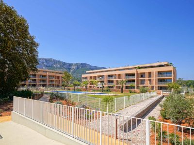 Vista exterior del residencial moderno con dos edificios de fachadas claras, piscina comunitaria y jardines. Al fondo, se aprecian las montañas bajo un cielo despejado.