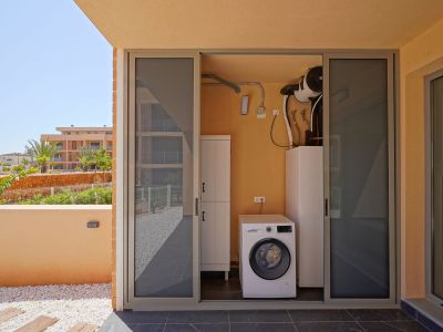 Zona de lavandería exterior con lavadora Bosch y sistema de aerotermia, accesible desde una terraza con suelo de gres, guijarros y vistas al jardín.