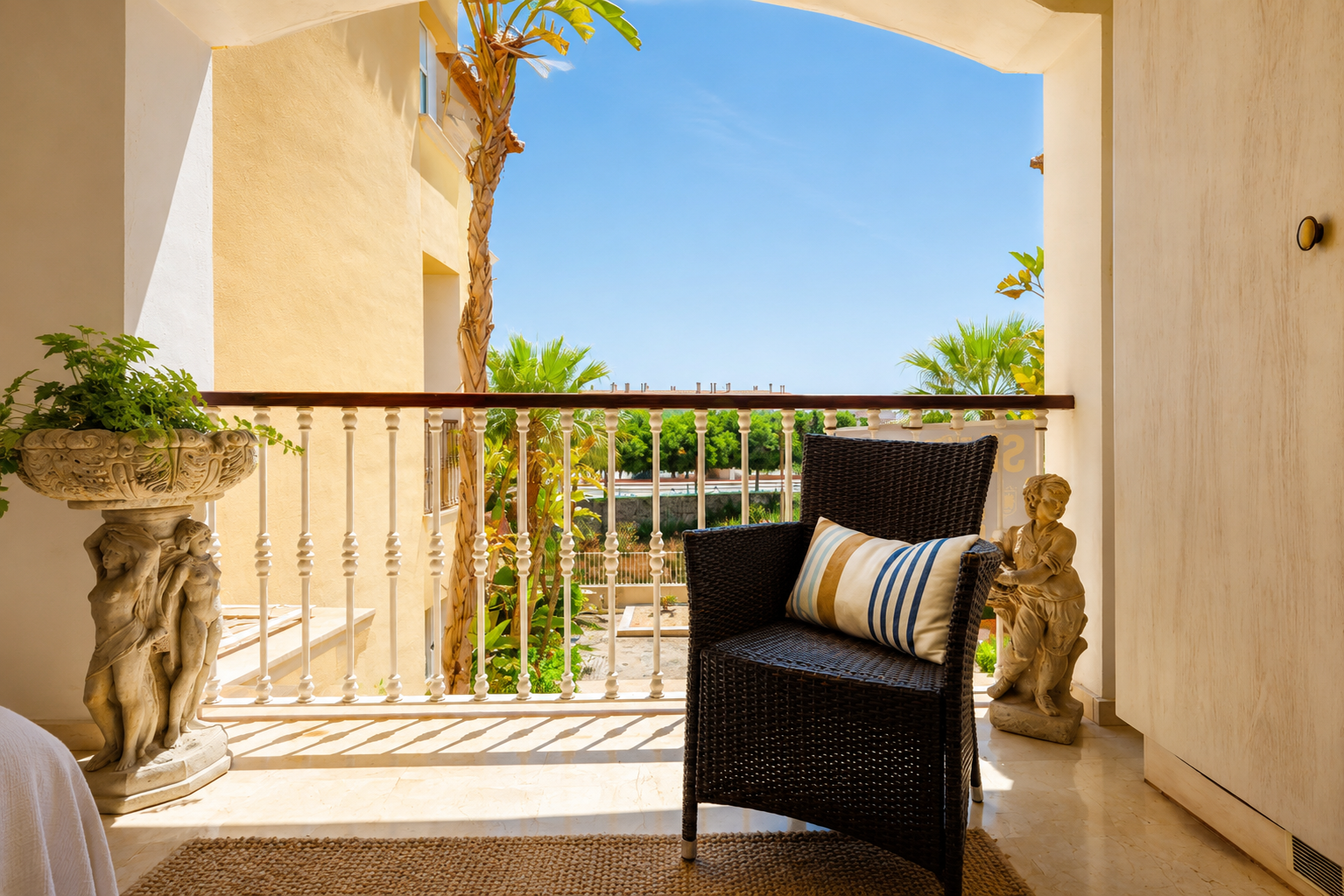 Agradable terraza privada con un sillón de mimbre oscuro, suelo de mármol claro y una balaustrada ornamentada. Incluye macetero y estatua de piedra, con vistas a palmeras y cielo azul.