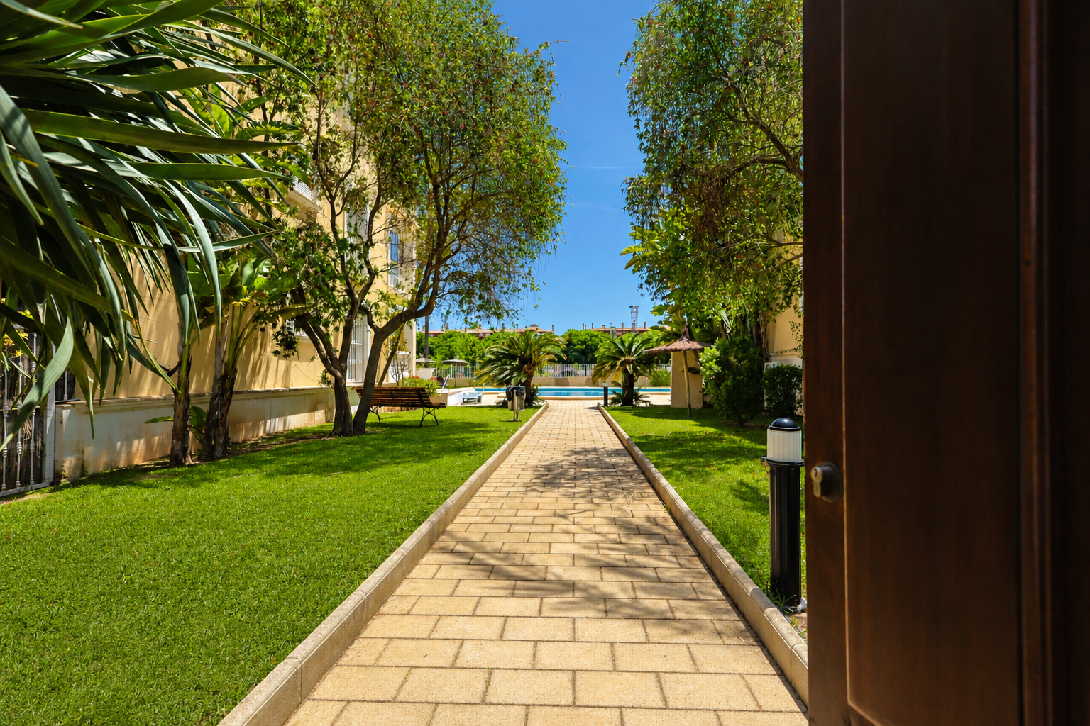 Camino pavimentado a través de los cuidados jardines comunitarios, revelando la piscina y las palmeras bajo un cielo azul. Vista desde una puerta abierta.
