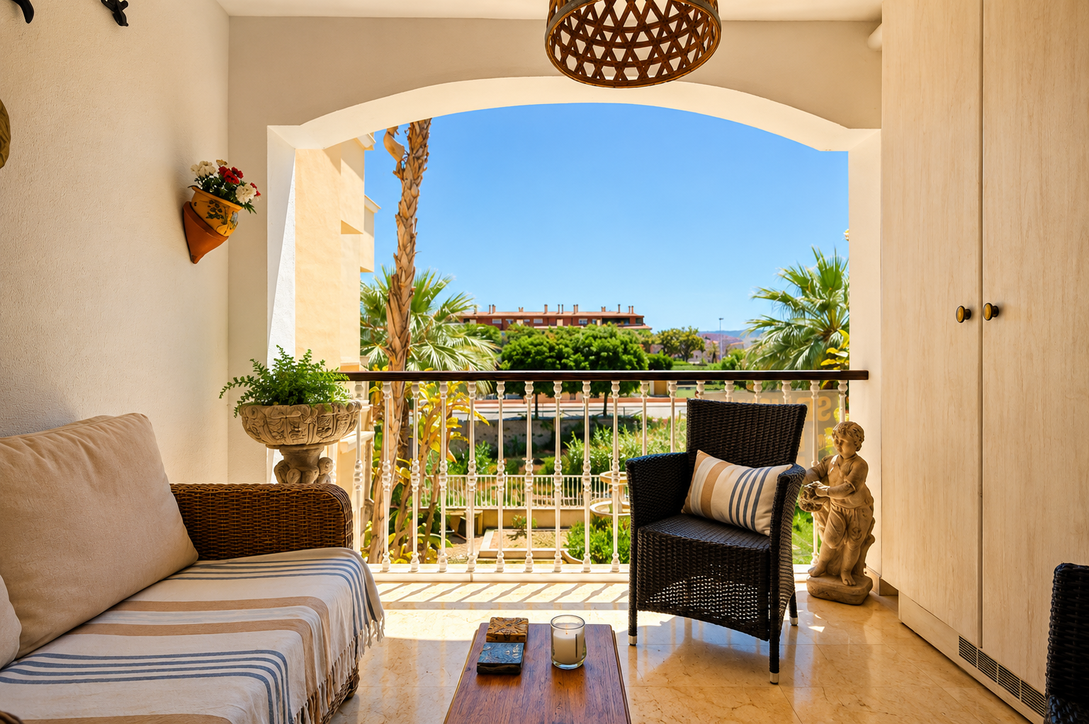 La terraza privada, con suelo de efecto mármol claro, ofrece sofá y sillón de mimbre. Un arco enmarca vistas a palmeras, jardines y edificios lejanos bajo cielo azul, con armarios de almacenamiento integrados.