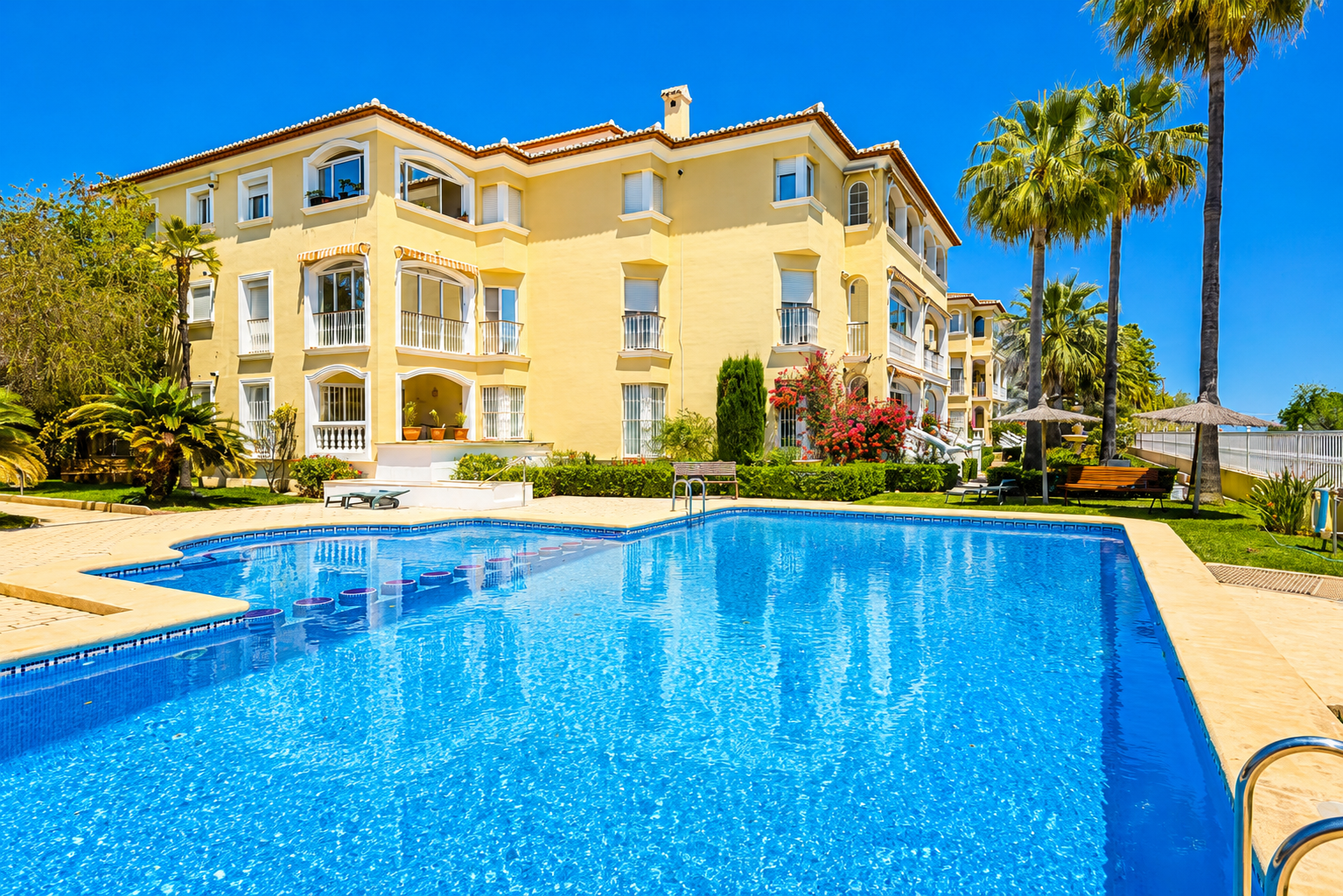La vibrante piscina comunitaria con mosaicos está rodeada por una terraza de baldosas beige y exuberantes jardines mediterráneos, con palmeras y el edificio amarillo bajo un cielo azul claro.