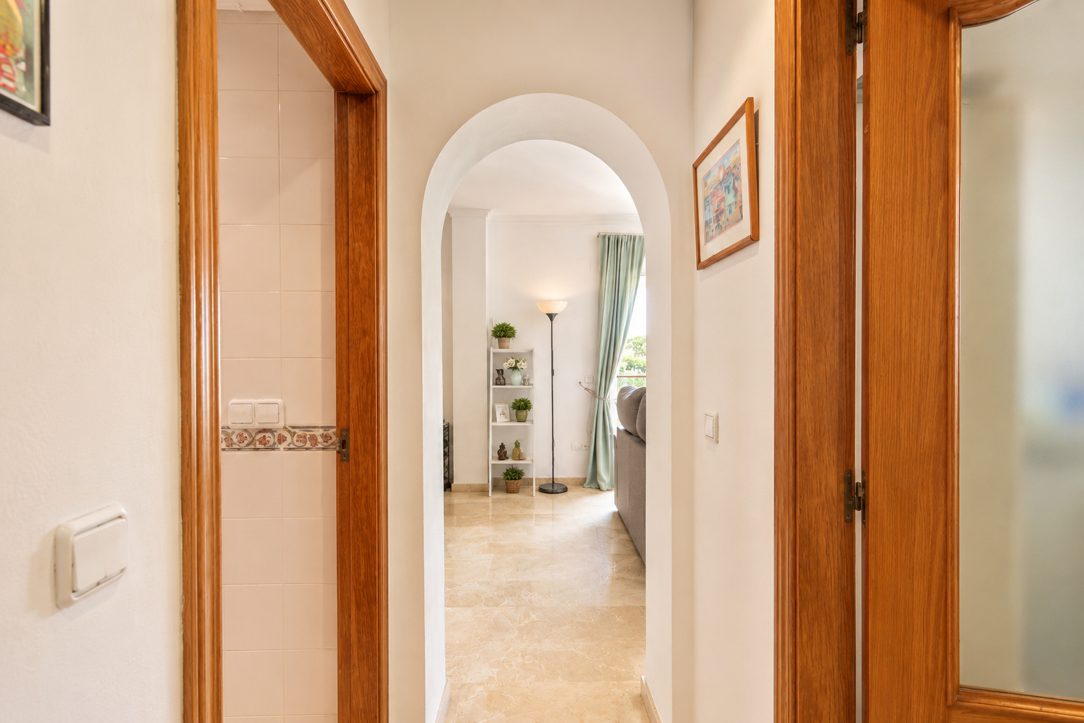 Pasillo con arco y suelo de mármol claro, conectando con sala de estar con ventana y cortinas azules. Marcos de madera flanquean el paso, uno revelando una pared alicatada.