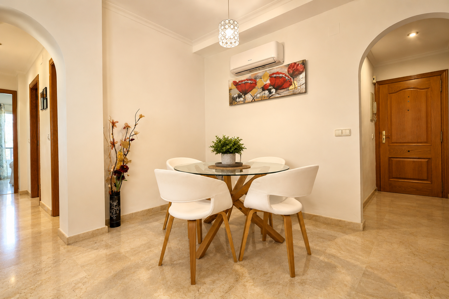 Zona de comedor compacta con mesa redonda de cristal y base de madera, cuatro sillas blancas y suelo de mármol claro. Un split de aire acondicionado y un cuadro floral decoran la pared.