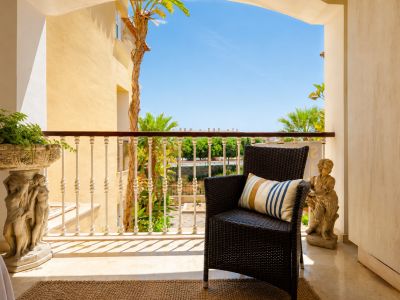 Agradable terraza privada con un sillón de mimbre oscuro, suelo de mármol claro y una balaustrada ornamentada. Incluye macetero y estatua de piedra, con vistas a palmeras y cielo azul.