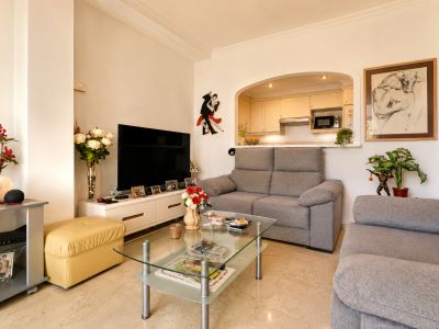 Amplio salón-comedor con sofás grises y mesa de centro de cristal, conectado a la cocina mediante un arco. Destaca el suelo de mármol claro y la iluminación natural.