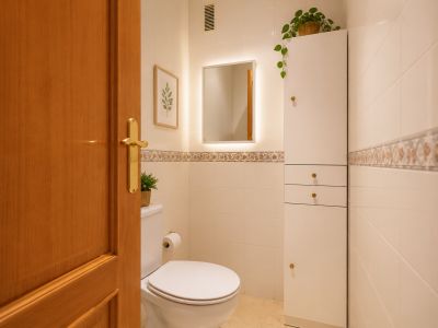 Baño funcional con paredes de azulejos crema y cenefa decorativa, inodoro blanco y espejo retroiluminado. Incluye un mueble alto blanco con tiradores dorados y puerta de madera.