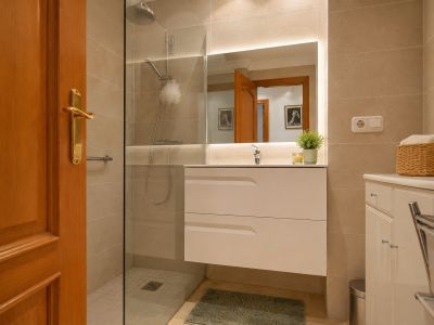 Baño moderno con lavabo flotante blanco y espejo iluminado. Incluye ducha de obra con mampara de cristal, paredes de azulejos beige y suelo de mármol claro.
