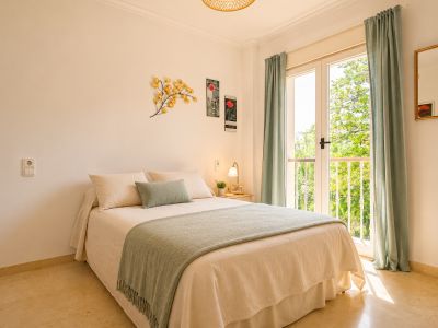 Dormitorio luminoso con cama doble, suelo de mármol claro y acceso directo a balcón con vistas a la vegetación. Cortinas verde menta y detalles decorativos en la pared.