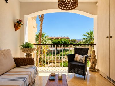 La terraza privada, con suelo de efecto mármol claro, ofrece sofá y sillón de mimbre. Un arco enmarca vistas a palmeras, jardines y edificios lejanos bajo cielo azul, con armarios de almacenamiento integrados.
