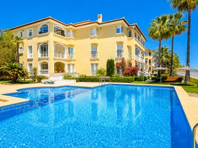 La vibrante piscina comunitaria con mosaicos está rodeada por una terraza de baldosas beige y exuberantes jardines mediterráneos, con palmeras y el edificio amarillo bajo un cielo azul claro.