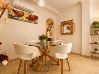 Luminoso comedor con mesa redonda de cristal y base de madera, sillas blancas, suelo de mármol claro y un espejo de madera tallada, complementado por una estantería decorativa.