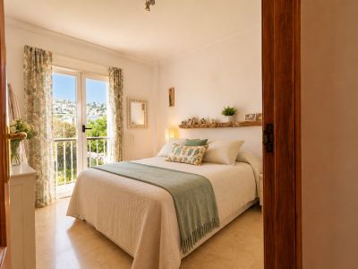 Luminoso dormitorio principal con acceso directo a balcón, ofreciendo vistas al pueblo y al mar. Destaca el suelo de mármol claro y la estantería de madera rústica sobre la cama.