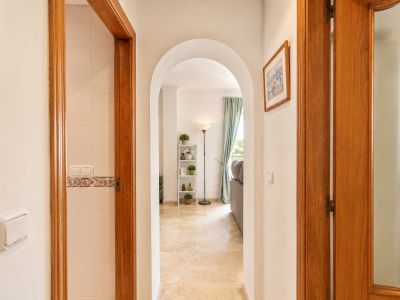Pasillo con arco y suelo de mármol claro, conectando con sala de estar con ventana y cortinas azules. Marcos de madera flanquean el paso, uno revelando una pared alicatada.